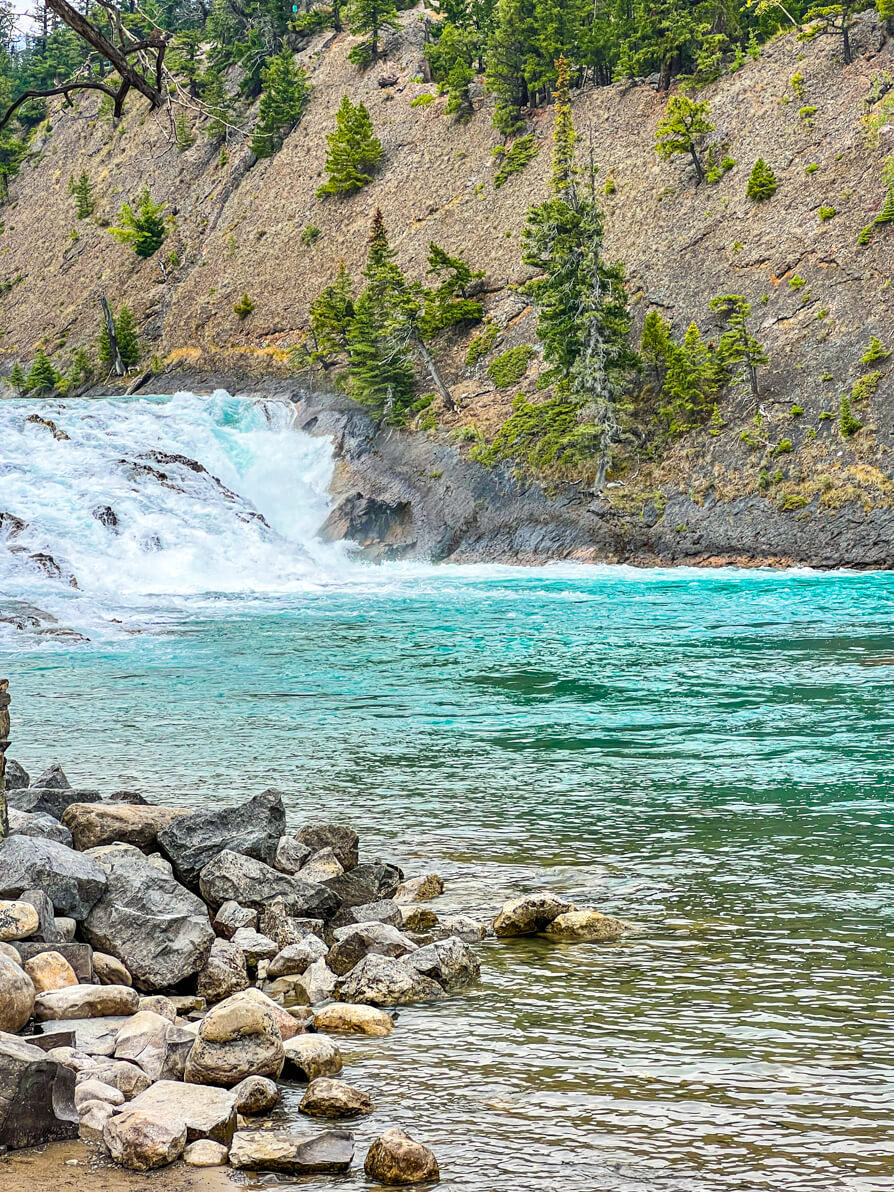 Image of Bow Falls in distance with rocks and green trees in forefront and turquoise blue Bow River in Banff Canada