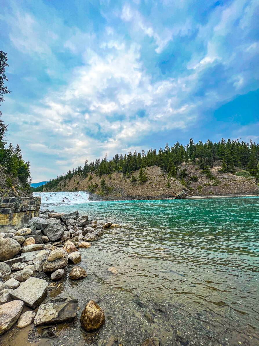 Image of Bow Falls in distance with rocks and green trees in forefront and turquoise blue Bow River flowing and blue sky with a few clouds in Banff Canada
