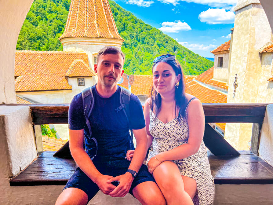 image of Shireen and Scott in front of Bran Castle courtyard with Carpathian Mountains and blue sky in background