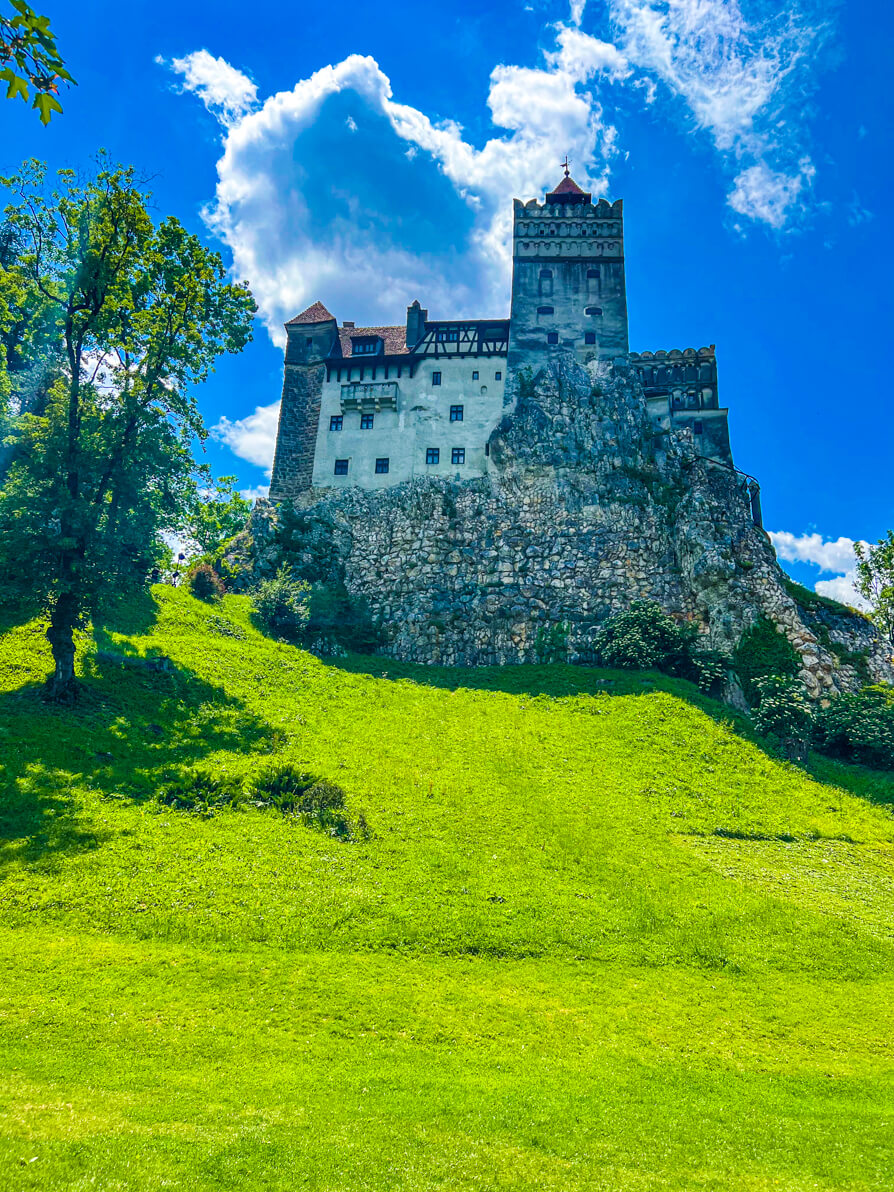 Image of Bran Castle Romania from the garden grounds. Grass in front and castle in upper back with blue sky behind and green trees to the left