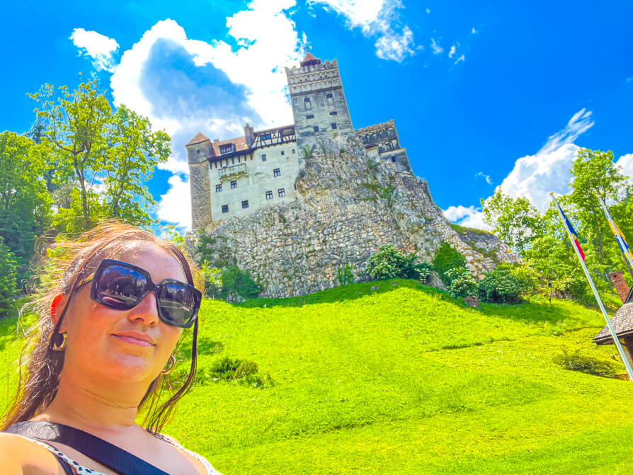 Image of Shireen in front of Bran Castle Romania from the garden grounds. Grass in front and castle in upper back with blue sky behind and green trees to the left