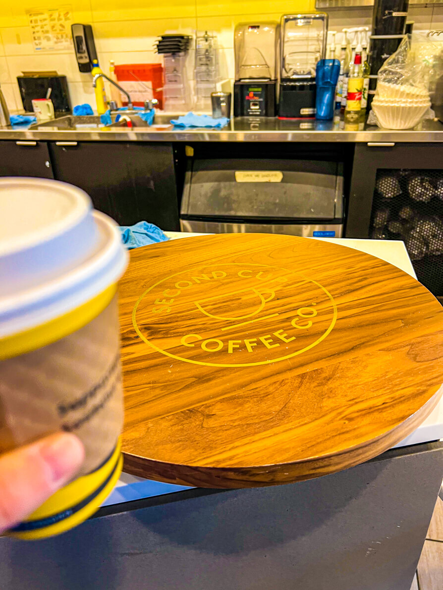 Image of Shireen's left hand holding a takeaway latte in front of second cup coffee sign at front desk in Banff
