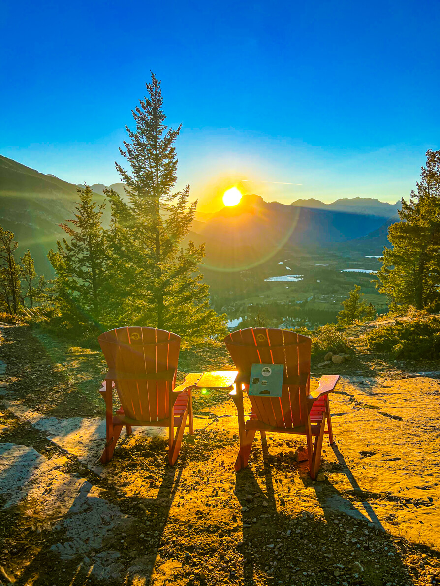 Image of two red deck chairs at the top of Tunnel Mountain trek with trees in centre, Rocky mountains in background and sun setting on the blue sky in Banff