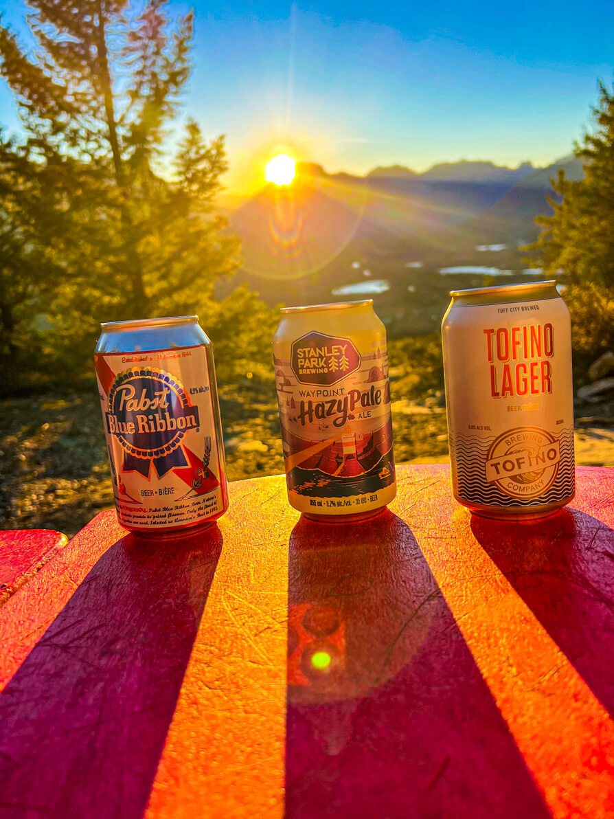 Image of three beer cans on Tunnel Mountain with rocky mountains and banff town in background as the sun sets