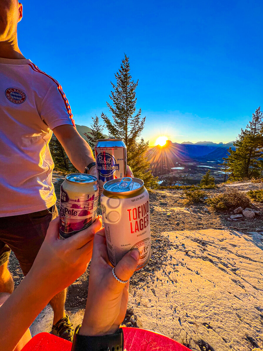 Image of Shireen and friends holding up beer cans on Tunnel Mountain with rocky mountains and banff town in background as the sun sets