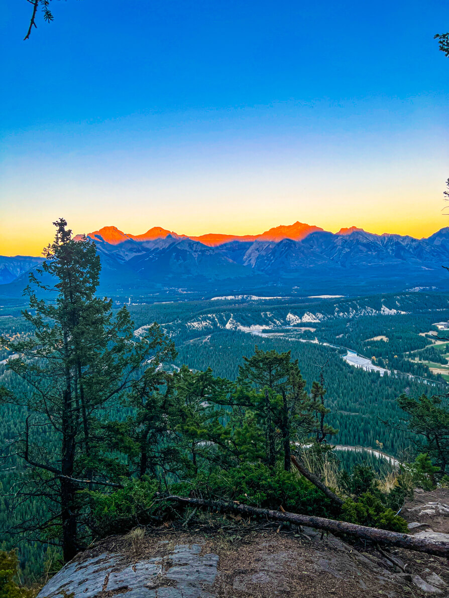 Image of rocky mountains with golden tips from the sun setting, river and golf course in centre of photo and green trees in forefront as seen from Tunnel Mountain