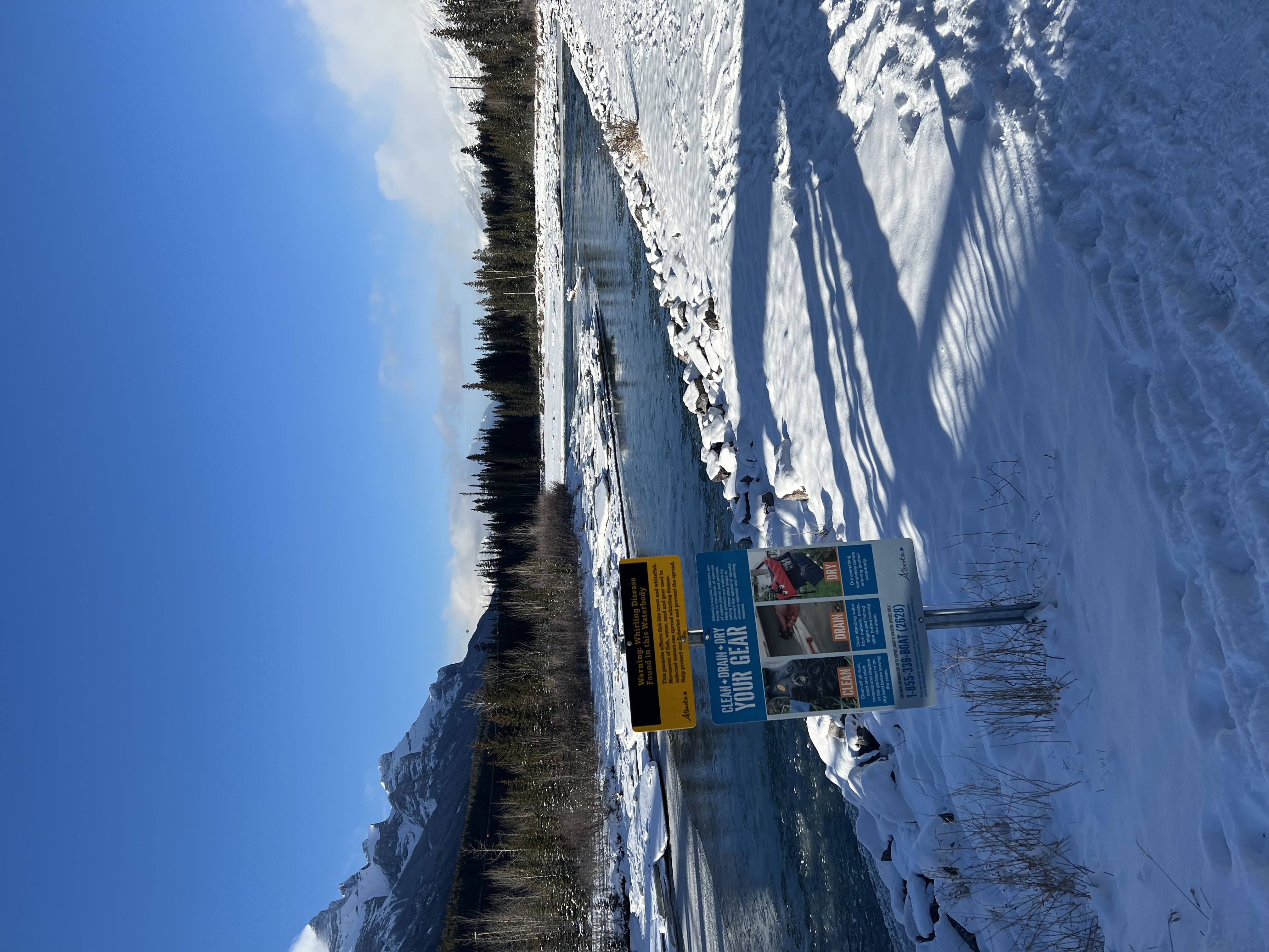 Start of the Canmore Engine Bridge. Image of the right hand side showing warning signs, snow on ground, river, trees and blue sky in background.