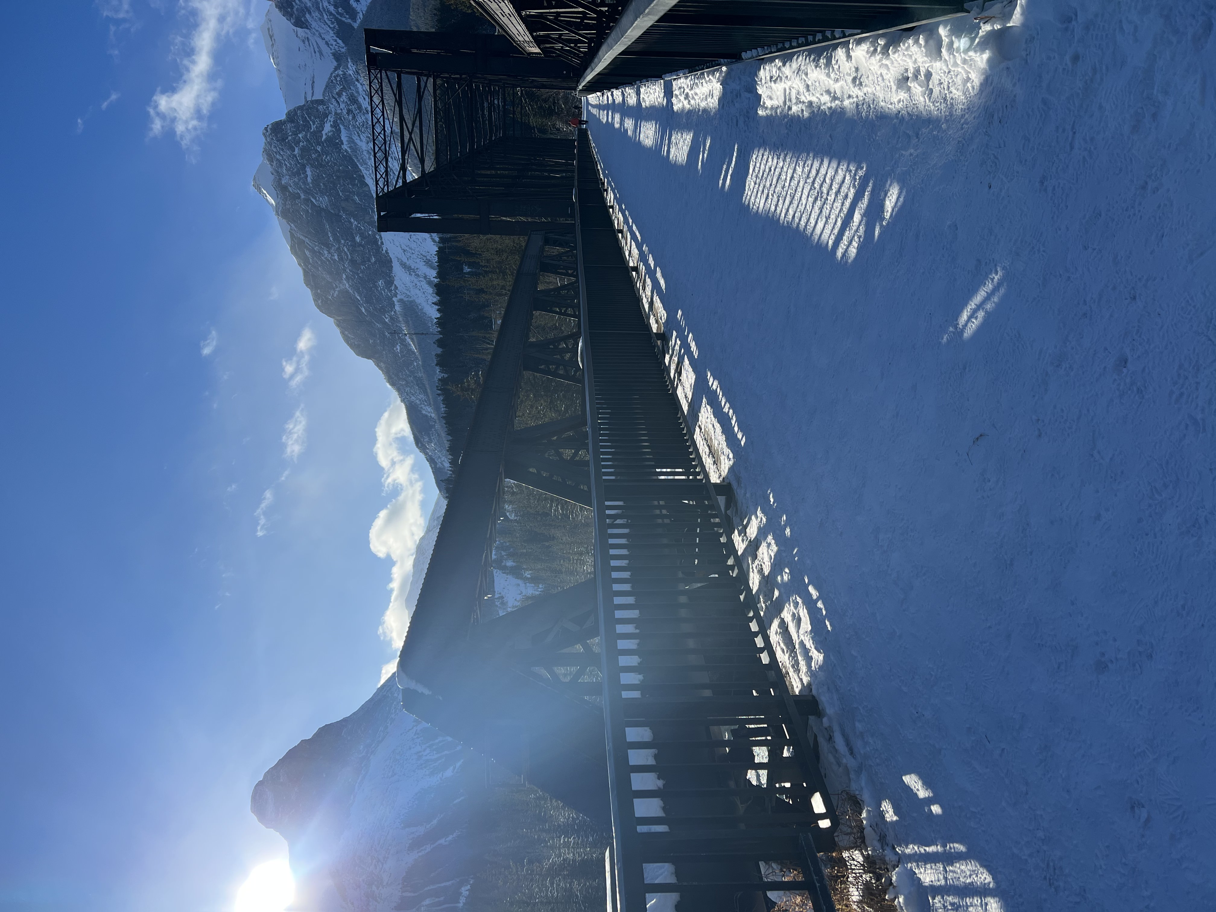 image of Canmore Engine Bridge in Canmore Alberta Canada. Filming Location for The Last of Us. Image taken from end of bridge with snow on ground, iron bridge either side of picture, mountains in background, blue sky and sun setting over far left mountain.
