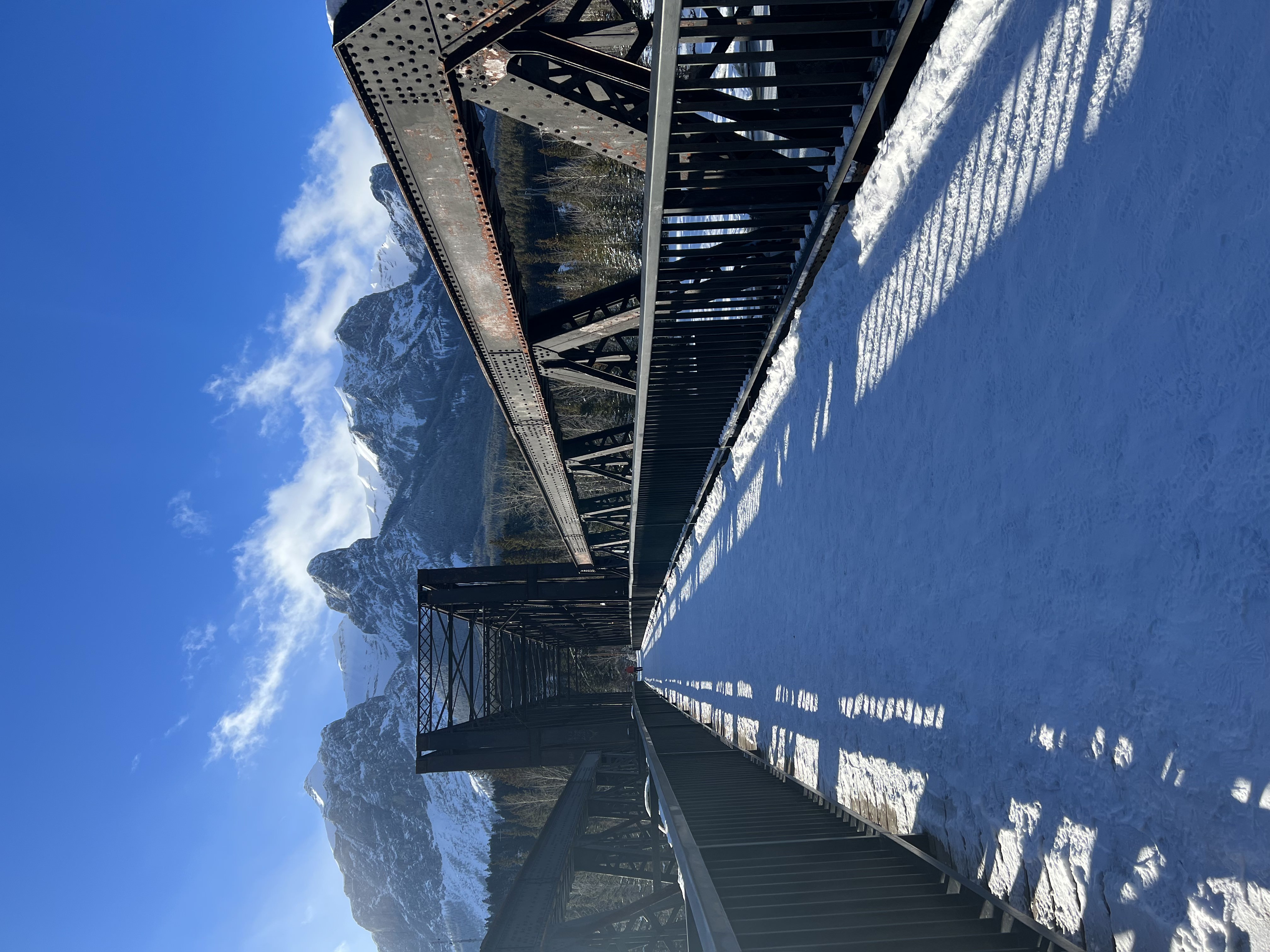 image of Canmore Engine Bridge in Canmore Alberta Canada. Filming Location for The Last of Us. Image taken from end of bridge with snow on ground, iron bridge either side of picture, mountains in background, blue sky.
