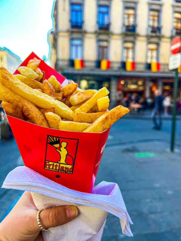 image of fritland Belgian fries. Shireen's left hand holding up Belgian Fries in a red cone with Belgium flags in the background displayed on a building in Brussels